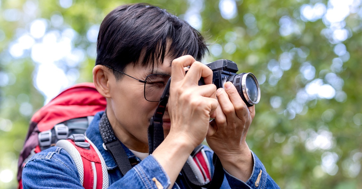 A man holding a professional camera up to his eye. He is outside, wearing a large red hiking backpack.