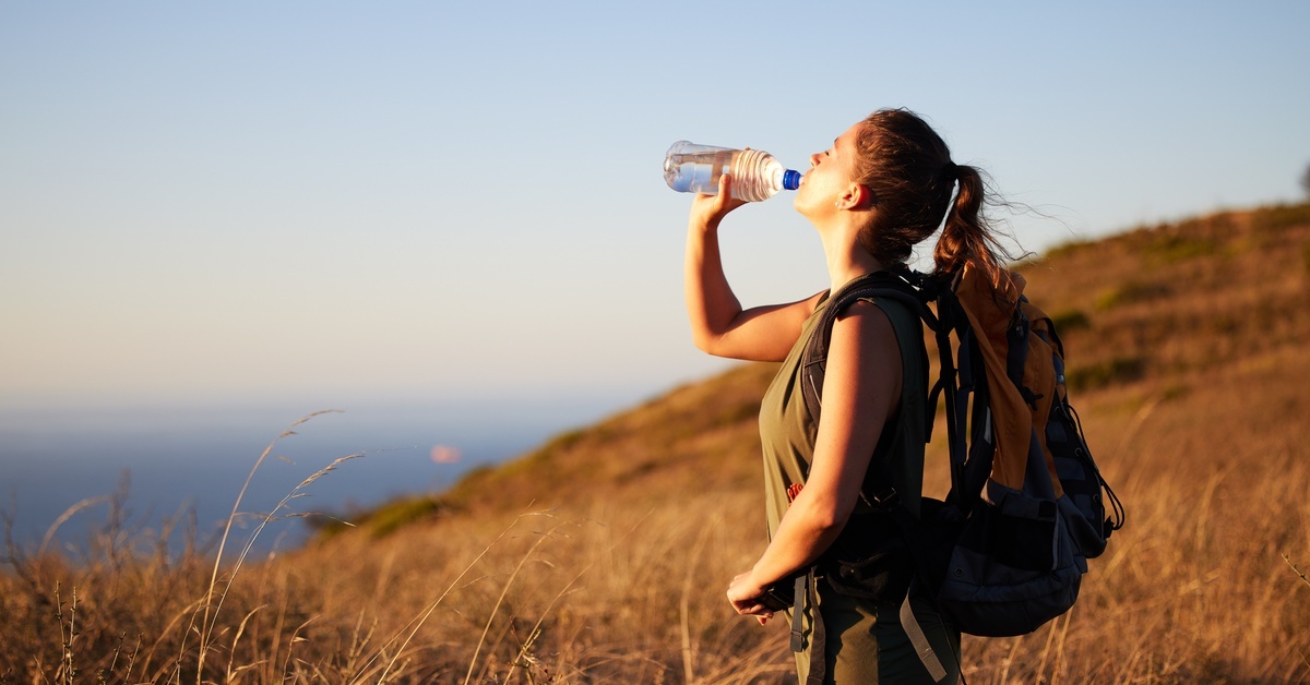 A woman wearing a hiking backpack and drinking from a plastic water bottle. She is in the middle of an expansive field.