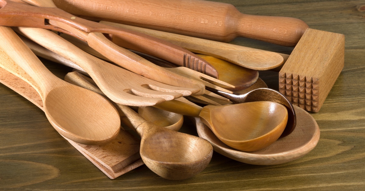 Wooden kitchen utensils lying on a dark brown surface. Large spoons, spatulas, and a rolling pin make up the pile.