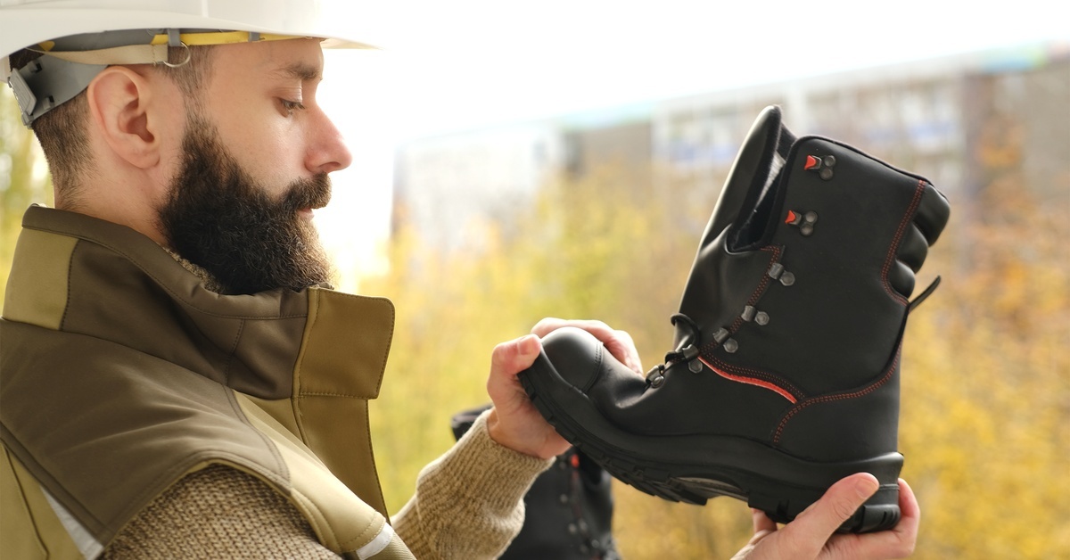 A man wearing a white hard hat inspects a black pair of steel-toe shoes. He bends the front of one shoe upwards.