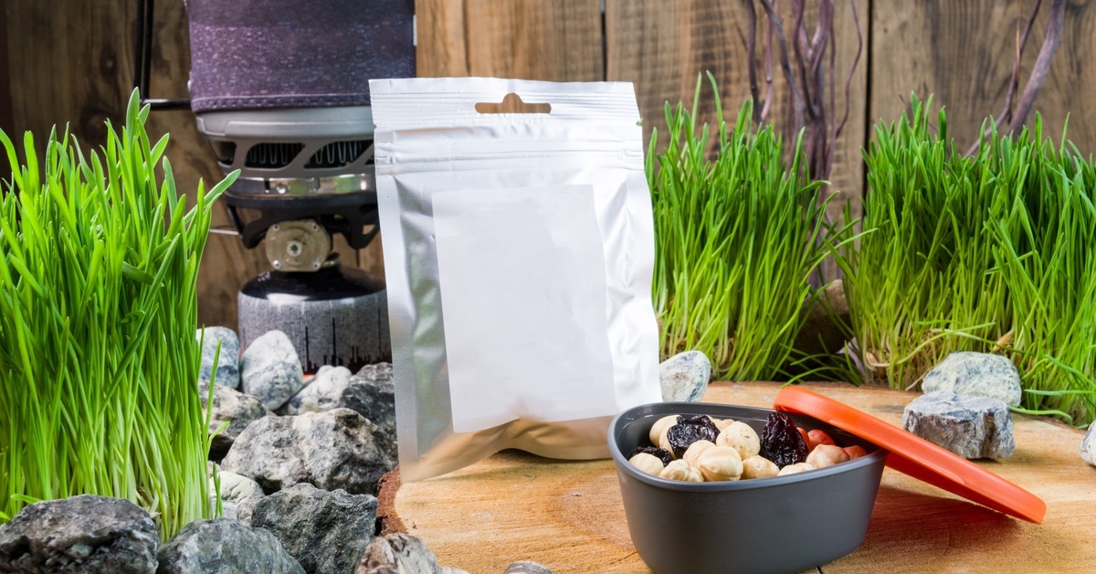 A tall grill sits on top of rocks behind a silver package and a food storage container laid out on a tree stump.