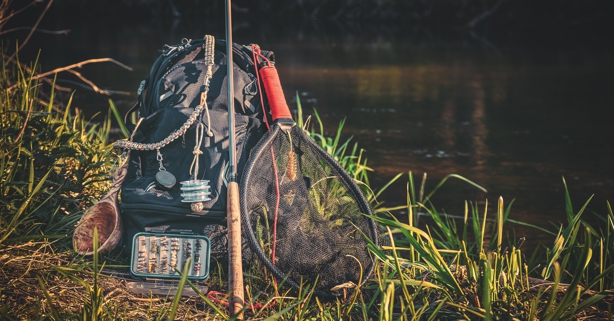 A fishing rod, net, and and other equipment are all piled onto a large backpack next to a riverbank.
