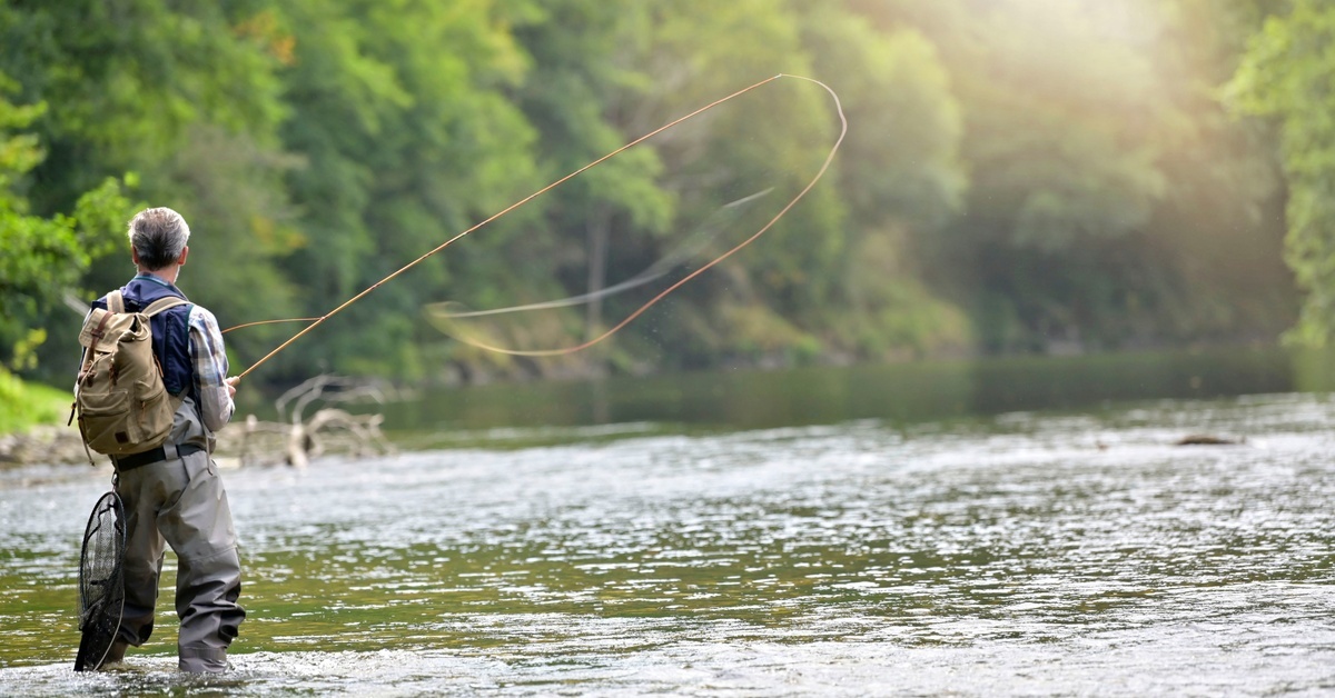 A person wearing a backpack stands in the middle of a lake while casting a fly fishing rod into the water.