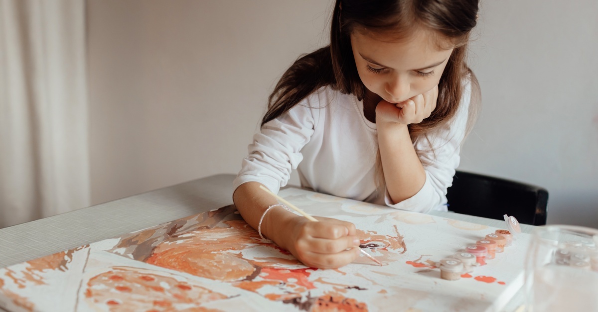 A little girl painting on a canvas with a small paintbrush. The painting is pink, orange, and brown in color.