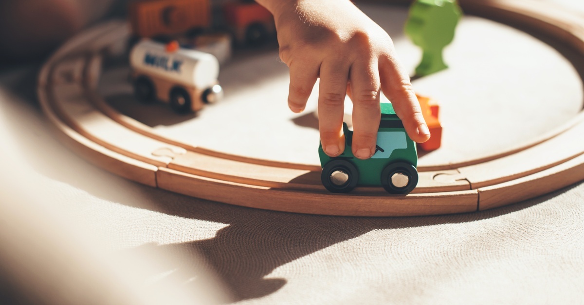 A small hand holding a toy train against a wooden track. The wooden track connects in a circle with other toy cars nearby.