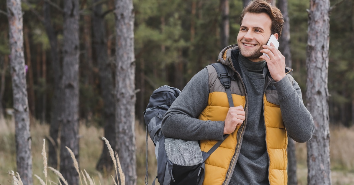 A man standing in the woods with his phone held up to his ear. He has a backpack slung over his shoulder.