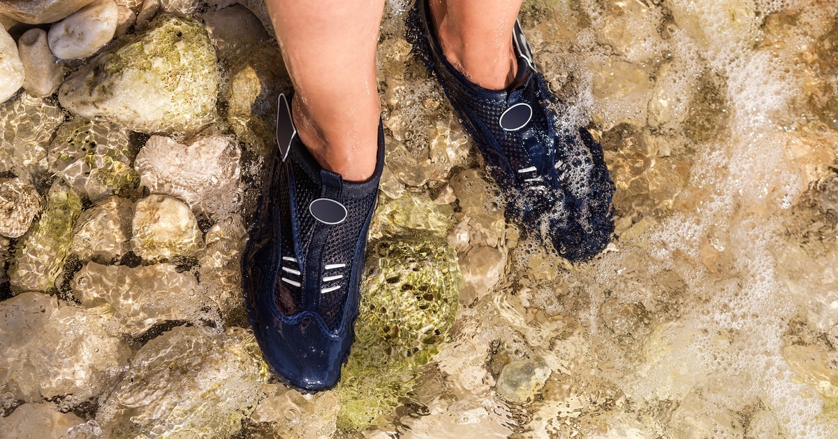 A close-up of a person wearing black water shoes outside. The shoes are submerged underwater at the shallow edge of a lake.