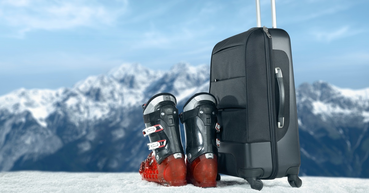 A pair of ski boots and a black suitcase sitting on snow-covered ground. There are ice-capped mountains in the background.