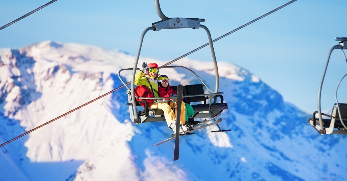 An adult and a child riding a ski lift together while wearing goggles and skis. One of them is waving.