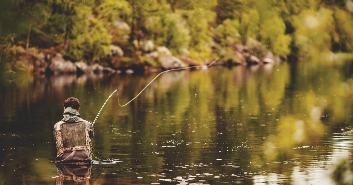 A man fly fishing in the middle of a lake with a fishing rod. He is standing waist-deep in the water.