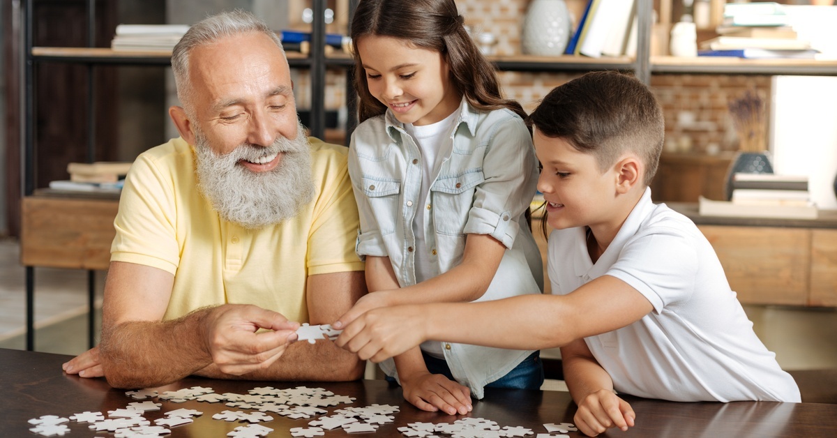 An older adult and two younger children solving a puzzle on a table in a living room. They are holding pieces in their hands.