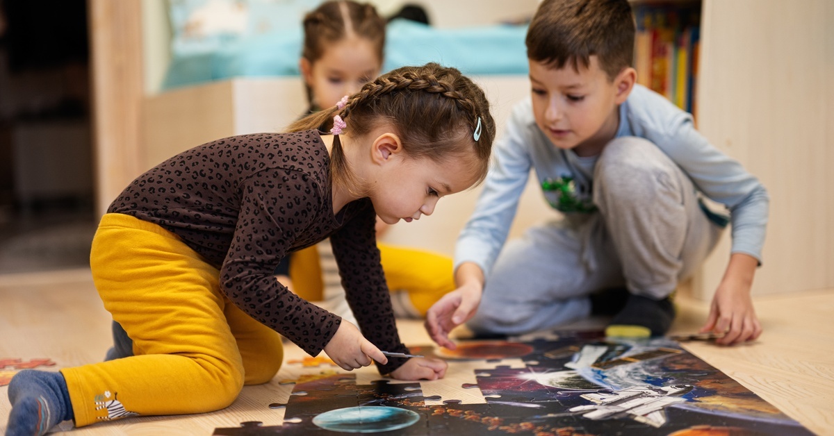Three kids solving a jigsaw puzzle on a wooden floor. The jigsaw puzzle depicts outer space, with planets and a spaceship.