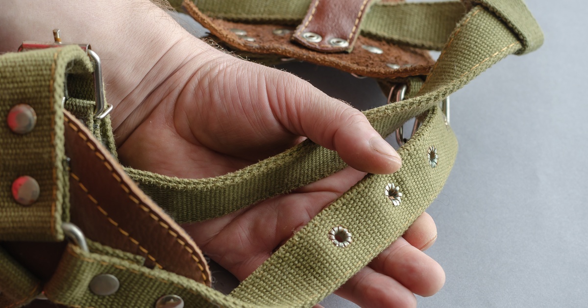 A person holding a dog harness in their hands against a gray background. The harness is made from a thick fabric and leather.