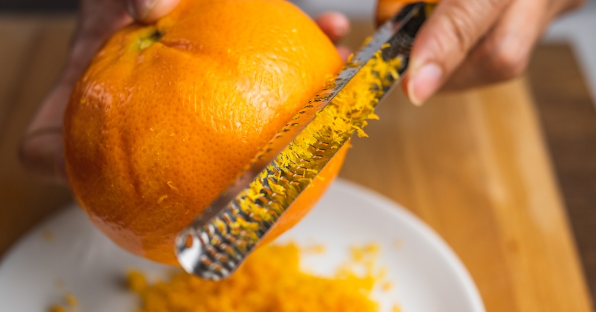 A person zesting a whole orange using a citrus zester. They collect the zest on a white plate on a wooden surface.